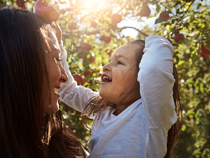 A mother holds her child in an apple orchard, with the child holding an apple in their hand--the Just One Health Impact Assessment Tool will offer a better way to measure progress and wellbeing